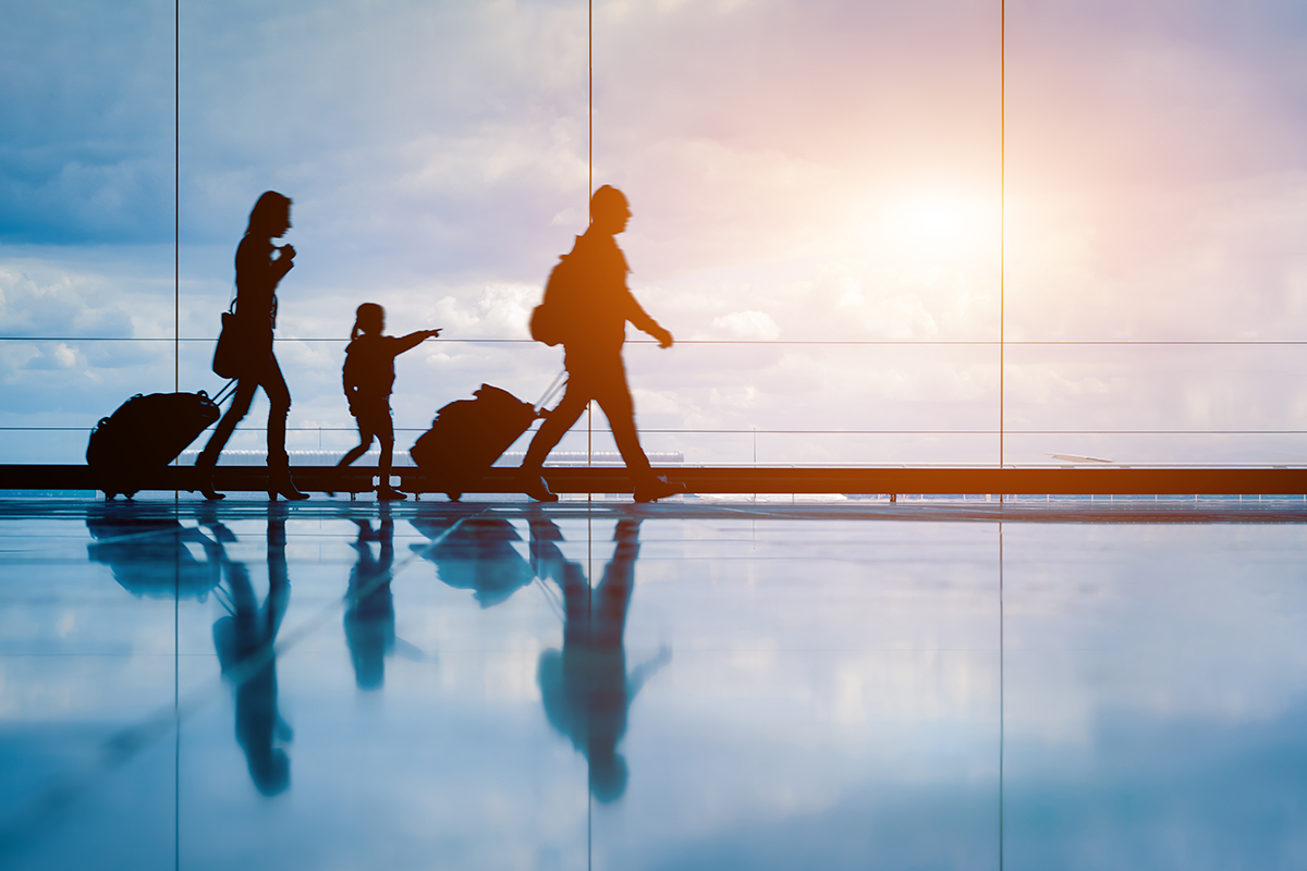 A mom, dad, and daughter with rolling suitcases walking next to airport of a sunny sky.